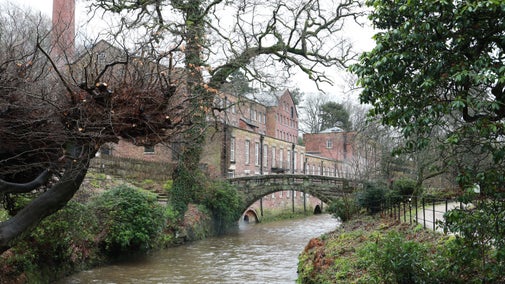 Long red brick mill building with stone bridge and brown river water below